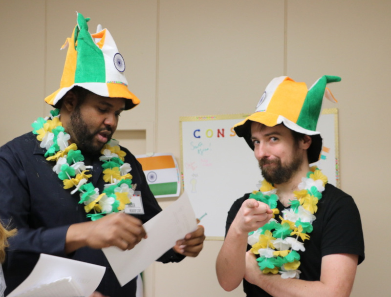 A photo of two men wearing hats and leis in with the colours of the Indian flag. There is a whitboard and an Indian flag in the background. One of the men is looking at a piece of paper, and the other is looking and pointing directly at the camera.