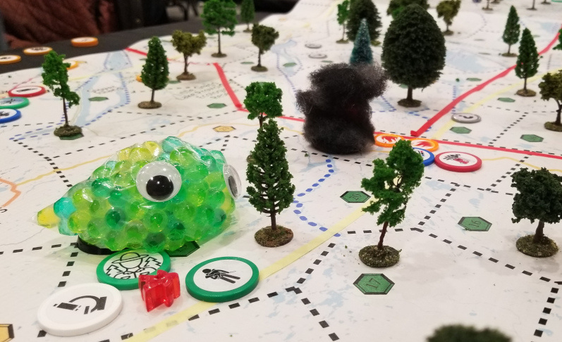 A photo of green blob with googley eyes sitting on a game board. The board has dotted and solid lines and several colours of game pieces. There are model trees sitting on the gameboard.