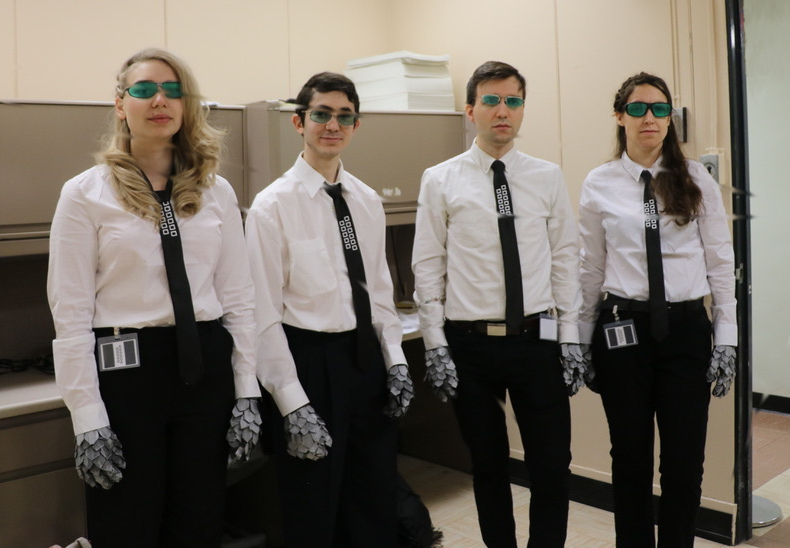 A photo showing a line of four people, dressed identically in green sunglasses, white shirts, black ties with silver hexagons, black pants, ID cards hanging off belt, and scaled silver gloves.