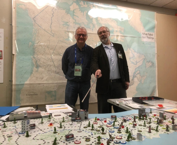 A photo of two men in front of a map of Canada. One of them has a pointer, and is pointing at some model buildings sitting on a game board in front of them.