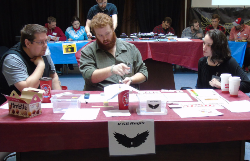 A photo with three peopole sitting around a red table. There are many pieces of paper and game cards on the table, and a sign for the table that reads 'ICSS Aegis' and has a wing icon. In the background, there are red and blue tables, with game pieces and people sitting at them.