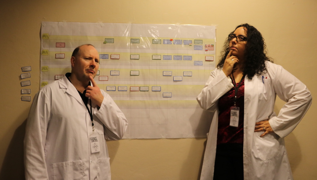 A photo of a man and a woman in labcoats, standing in front of an organizational chart. They both have their index fingers pointing at their chins, and they look puzzled.